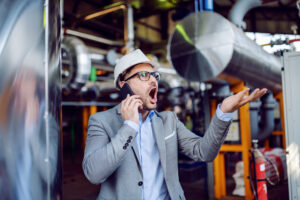 Angry businessman in suit and with protective helmet on head yelling over smart phone while standing in power plant.